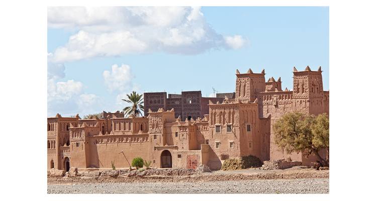Fortaleza de adobe estilo kasbah con torres almenadas y palmera contra un cielo azul.