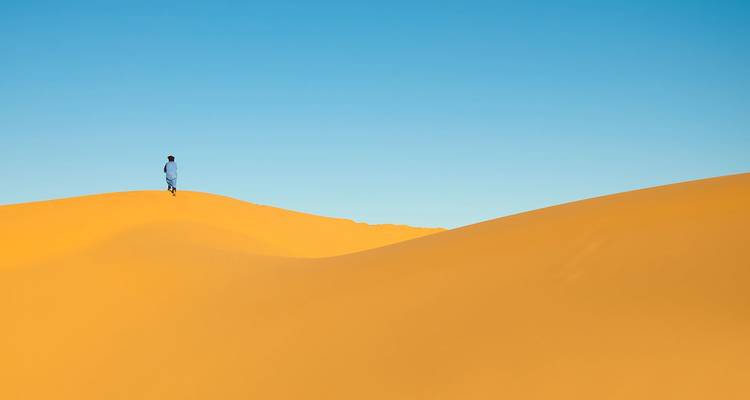 Scène minimaliste d'une silhouette solitaire se tenant au sommet d'une dune de sable doré et lisse sur un ciel bleu dégagé