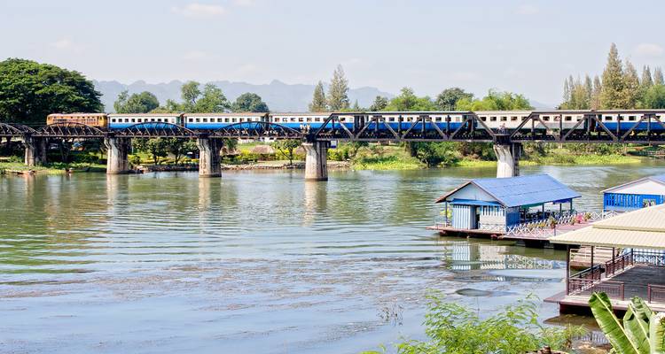 Trein steekt de historische stalen brug over een rustige rivier over met verre groene heuvels in Kanchanaburi