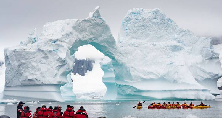 Kajakkers peddelen langs een gigantische gebeeldhouwde ijsbergboog terwijl een andere groep vanaf zodiacs toekijkt