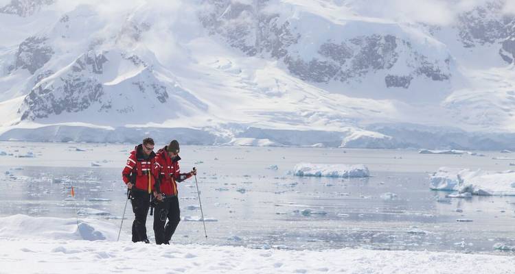 Twee ontdekkingsreizigers op ski's lopen over verse sneeuw naast een bevroren baai bezaaid met ijsschotsen