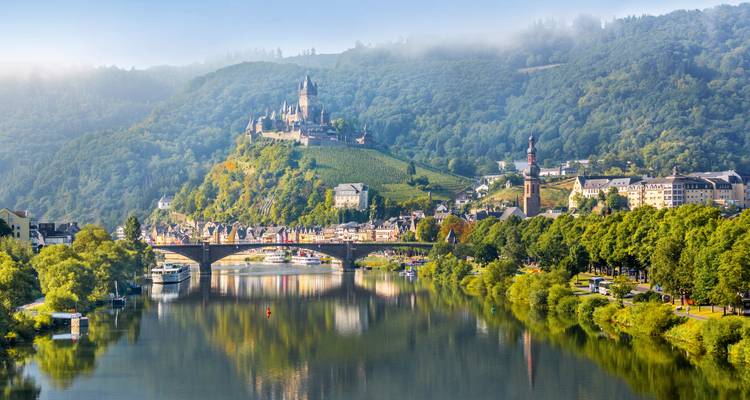 Die Burg Cochem krönt einen weinbergbedeckten Hügel, der sich an einem nebligen Morgen im ruhigen Wasser der Mosel spiegelt.