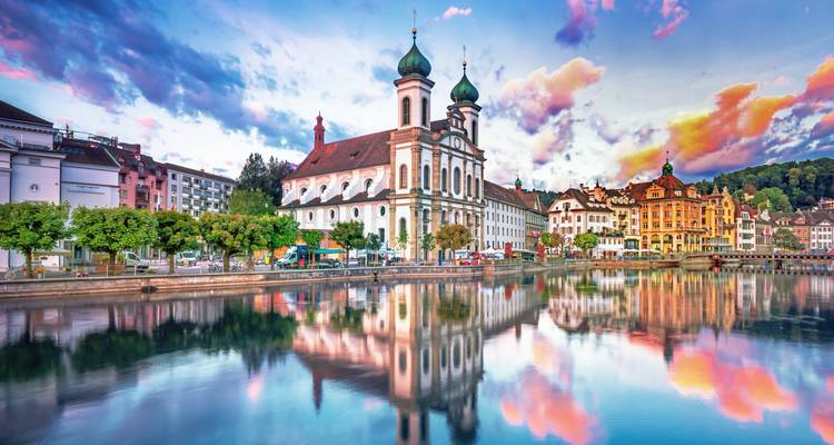 Barocke Jesuitenkirche und bunte Ufergebäude spiegeln sich bei Abenddämmerung perfekt in Luzerns Reuss wider.