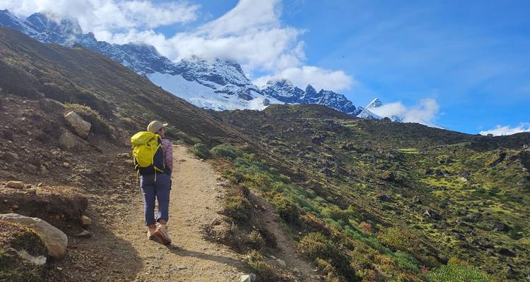 Backpacker met gele rugzak wandelt over een onverharde route richting besneeuwde Himalaya-toppen onder een stralend blauwe hemel