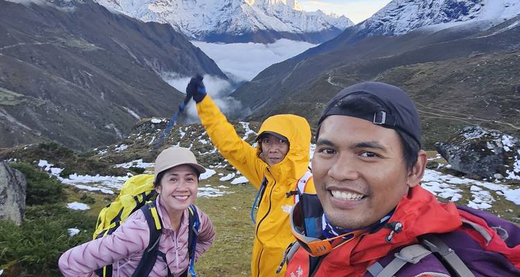 Drie trekkers die glimlachen voor een selfie op een bergpad met besneeuwde Himalaya-toppen en een met wolken gevulde vallei achter hen.
