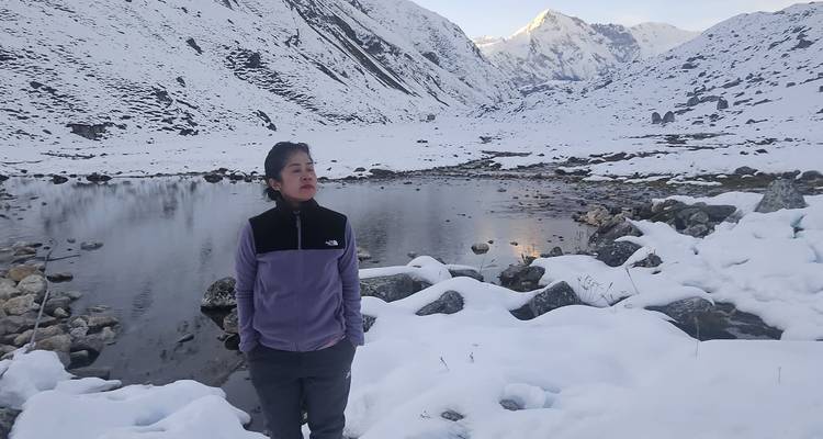 Een vrouw staat naast een sereen, gedeeltelijk bevroren alpenmeer omringd door besneeuwde bergen in de schemering.
