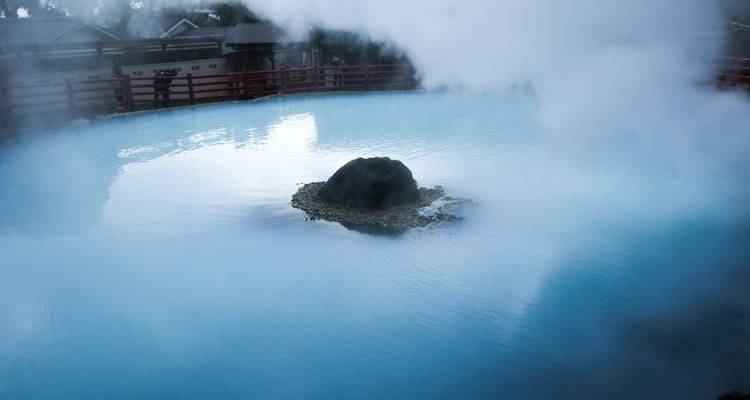 Bassin d'eau chaude turquoise fumant avec de la vapeur s'élevant autour d'une roche centrale.