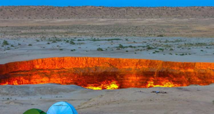 Vue nocturne du cratère ardent de Darvaza rougeoyant orange avec des tentes installées à proximité sur le sol du désert.