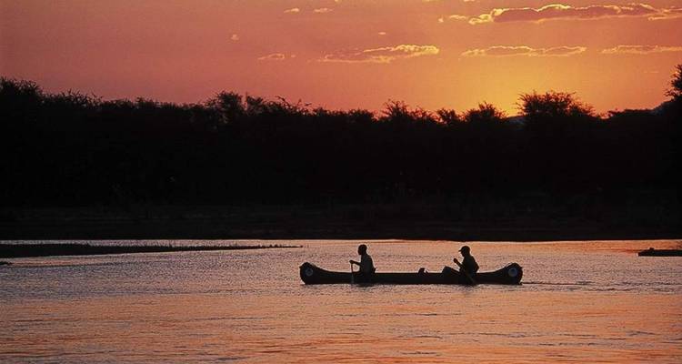 Silhouettiertes Kanu mit zwei Paddlern gleitet bei leuchtend orangenem Sonnenuntergang über einen ruhigen Fluss.