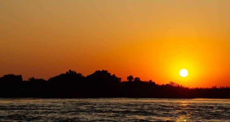 Strahlend orangene Sonne tief am Horizont über fließenden Flusswassern in der Dämmerung.