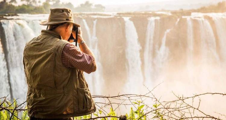 Ein Fotograf in Safari-Kleidung fotografiert die donnernden Victoriafälle von einem eingezäunten Aussichtspunkt aus.