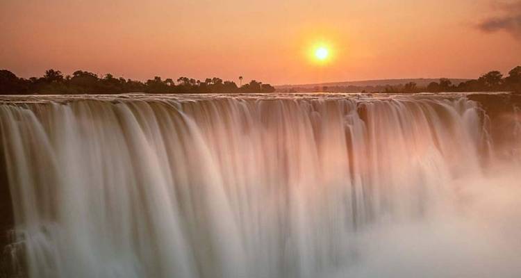 Die Sonne geht über den Viktoriafällen unter, goldenes Licht erhellt das glatt fließende Wasser und den Nebel.