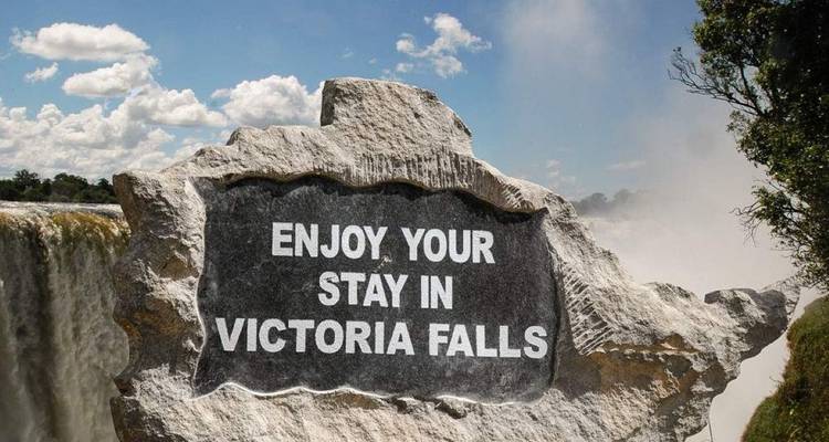 Steinschild mit der Gravur 'Genießen Sie Ihren Aufenthalt in Victoria Falls' neben dem nebligen Rand des Wasserfalls.