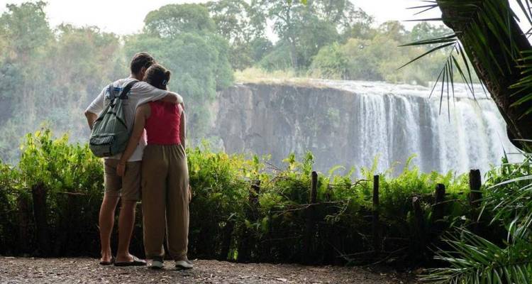 Ein Paar umarmt sich, während es die Victoriafälle durch üppiges Grün von einem schattigen Aussichtspunkt aus bewundert.