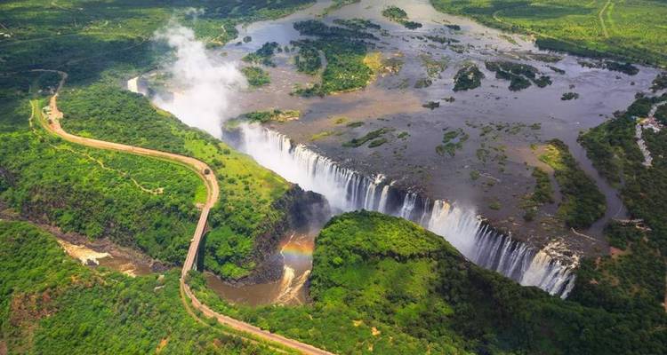 Luftaufnahme der Victoriafälle und des sich windenden Sambesi-Flusses mit Regenbogen und der umliegenden grünen Landschaft.