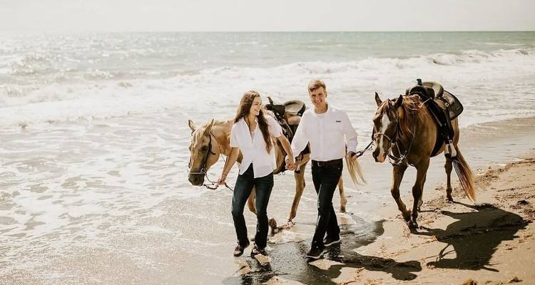 Ein junges Paar führt zwei Pferde entlang einer sonnigen Küstenlinie mit Wellen, die in der Nähe brechen