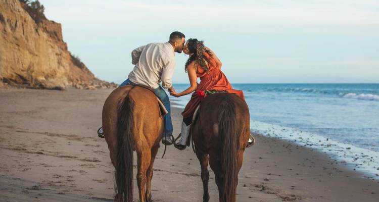 Paar zu Pferd teilt einen Kuss während des Reitens entlang eines abgelegenen Strandes unter Küstenklippen