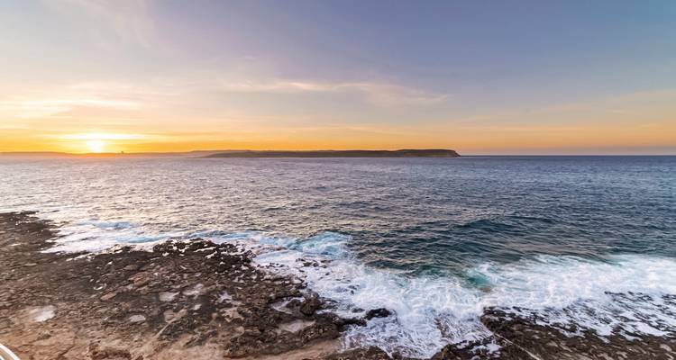 Felsige Küste mit brechenden Wellen unter einem farbenfrohen Abendhimmel mit Blick auf eine entfernte Landzunge.