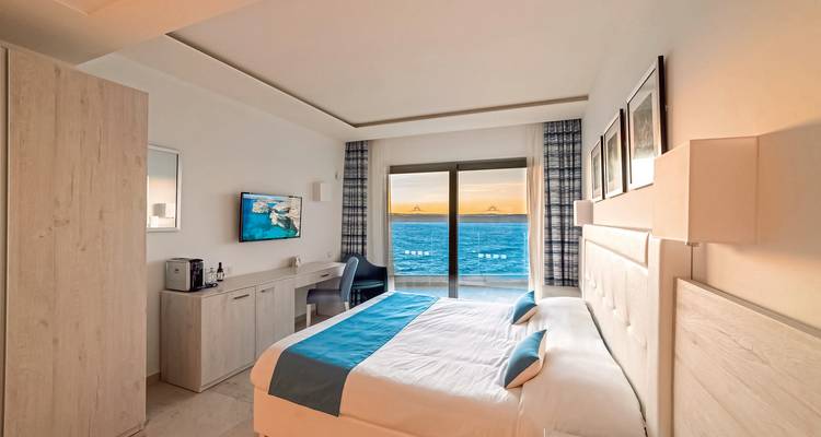 Modernes Hotelzimmer mit türkisfarbenen Akzenten mit Blick auf einen Balkon und das offene Meer bei Sonnenuntergang.