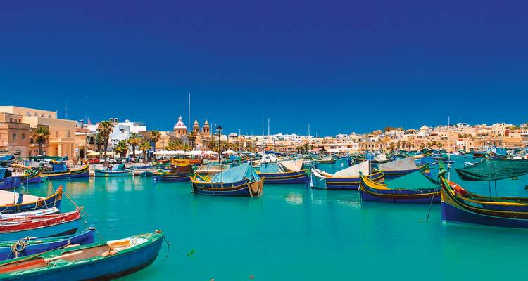 Helle Szene des Hafens von Marsaxlokk voller bunter traditioneller Luzzu-Fischerboote auf türkisfarbenem Wasser.