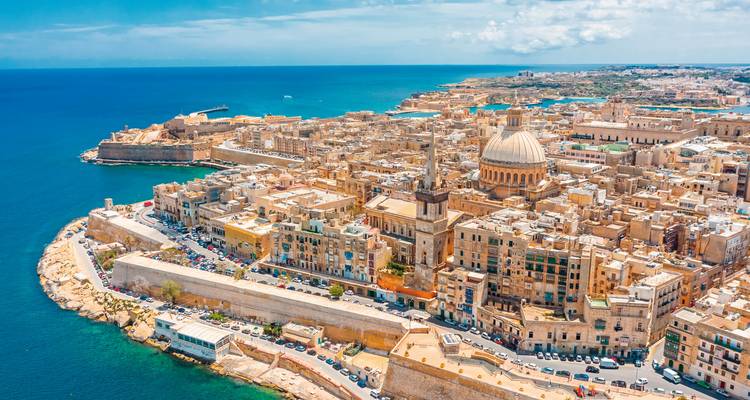 Weite Luftaufnahme der Halbinsel Valletta mit einer Kuppelkathedrale, die sich über honigfarbene Gebäude und tiefblaues Meer erhebt.
