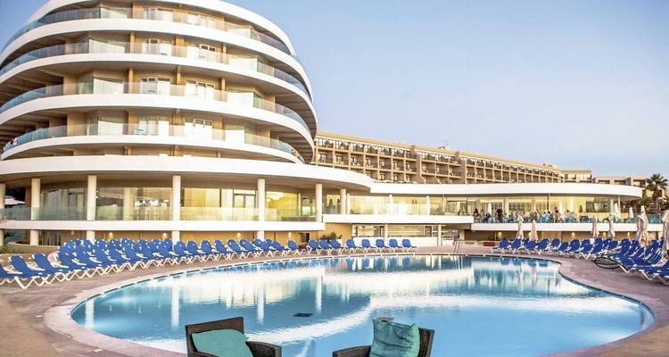 Modernes geschwungenes Hotelgebäude spiegelt sich in einem runden Swimmingpool mit Reihen blauer Liegestühle wider.