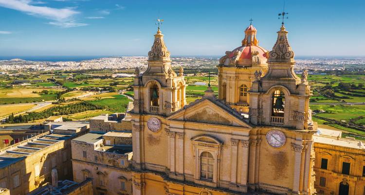 Luftaufnahme der barocken Kathedraltürme von Mdina, die sich über Maltas ländliche Landschaft unter blauem Himmel erheben.