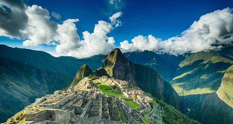 Panoramic view of Machu Picchu ruins beneath steep green peaks and a bright blue sky with scattered clouds.