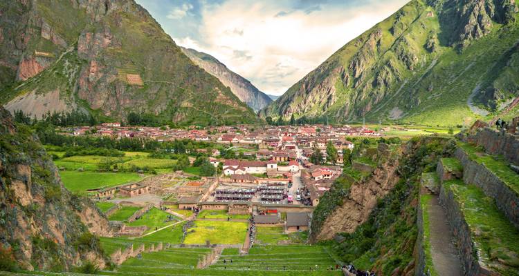 Elevated look down onto the town of Ollantaytambo framed by steep green Andean mountains and terraced ruins.