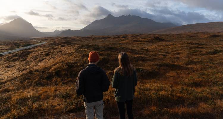 Deux voyageurs se tiennent sur une lande de bruyère, absorbant de vastes panoramas montagneux dans une lumière dorée