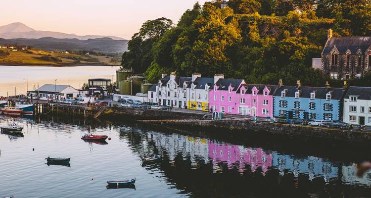 Reihe von bunt bemalten Hafenhäusern, die sich zur goldenen Stunde im ruhigen Wasser von Portree spiegeln.