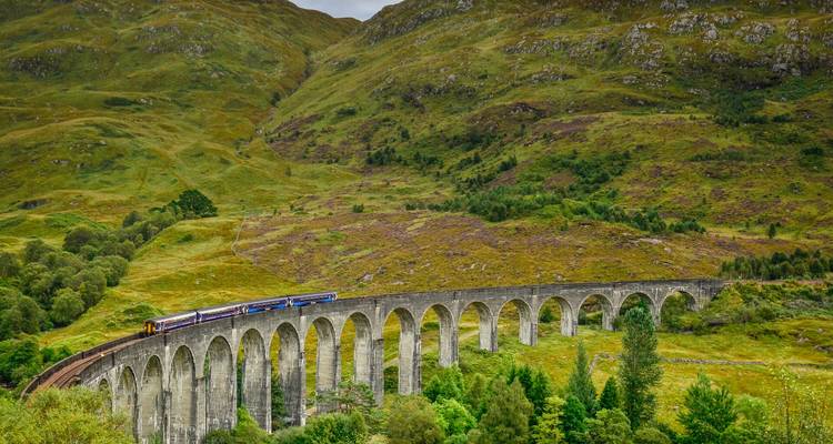 Un train à vapeur historique traverse le majestueux viaduc de Glenfinnan au milieu de collines verdoyantes ondulantes.