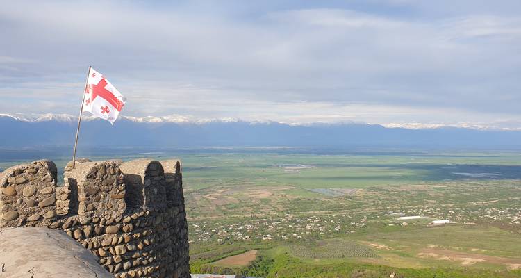 Steinerner Festungsturm mit georgischer Flagge überblickt weite grüne Ebenen und schneebedeckte Kaukasus-Berge.