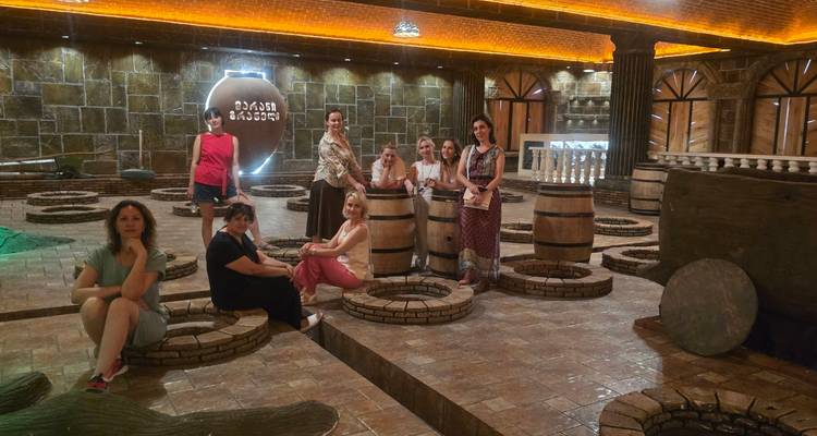 Gruppe von Frauen, die in einer weinkellerartigen Halle mit Backsteinwänden, Holzfässern und kreisförmigen Gruben posieren.