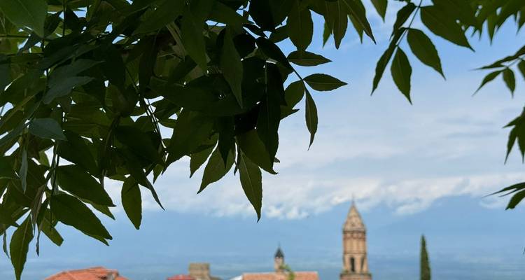 Kirchturm und rote Ziegeldächer schauen durch belaubte Äste hervor mit schneebedeckten Kaukasus-Bergen in der Ferne.