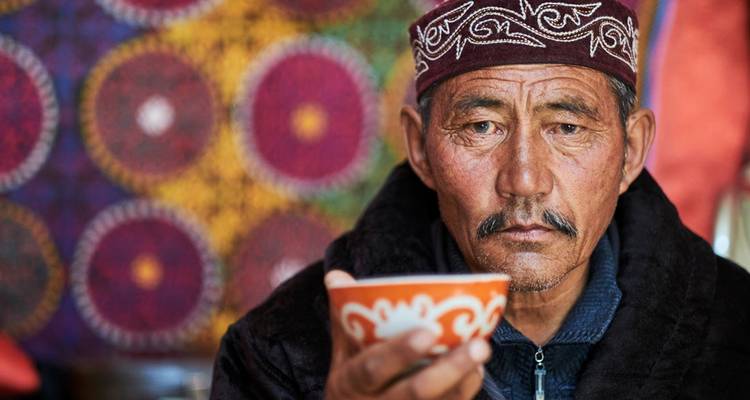 Portrait d'un homme d'Asie centrale âgé portant un chapeau traditionnel et tenant un bol à thé à motifs avec des textiles colorés derrière lui.