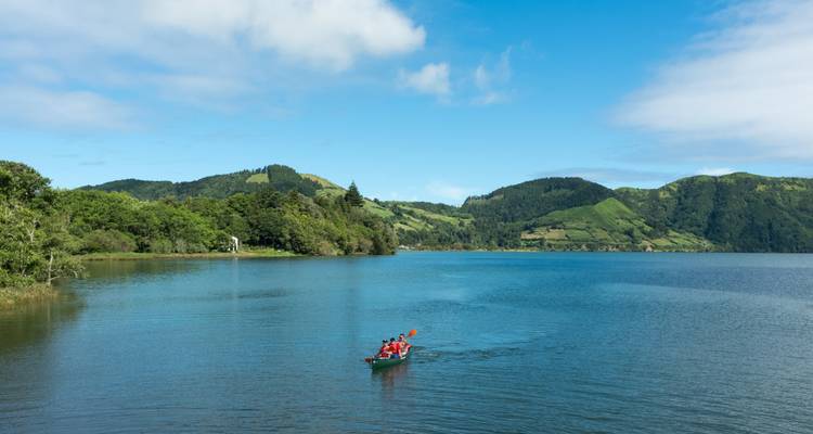 Tres personas reman en una canoa roja sobre un lago volcánico azul sereno rodeado de colinas boscosas.