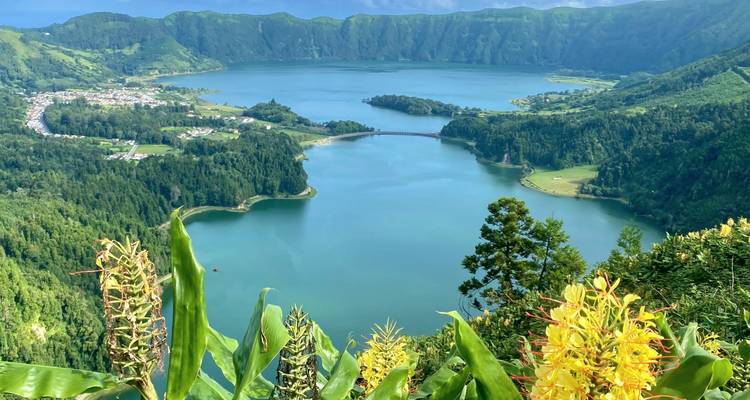 Vista elevada de la caldera de Sete Cidades con lago azul vívido, pueblo y laderas volcánicas exuberantes enmarcadas por flores.