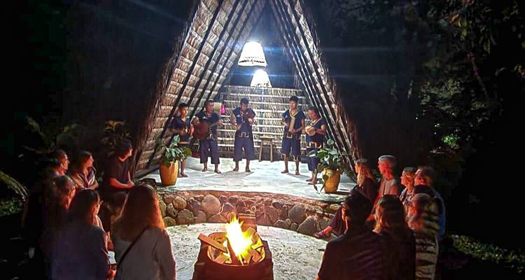 Spectacle culturel nocturne autour d'un feu de camp à l'extérieur d'une structure en A au toit de chaume devant un public qui regarde.
