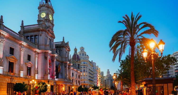 Abendszene eines lebhaften Platzes mit beleuchteten historischen Gebäuden, Palmen und Straßenlaternen.