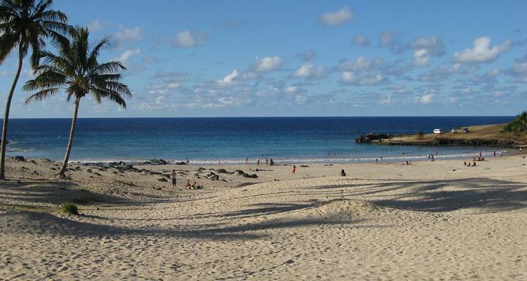 Playa de arena blanca con visitantes dispersos, palmeras y mar turquesa en la Isla de Pascua.