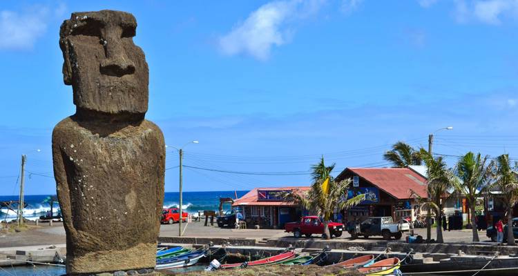 Estatua moai solitaria con vista a un puerto con botes coloridos y pueblo costero en la Isla de Pascua.