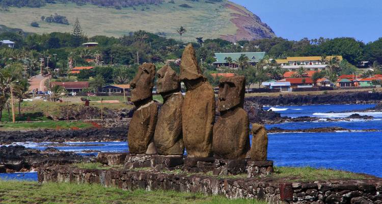 Vista trasera de cinco estatuas moái mirando hacia el mar con pueblo costero y colinas al fondo.
