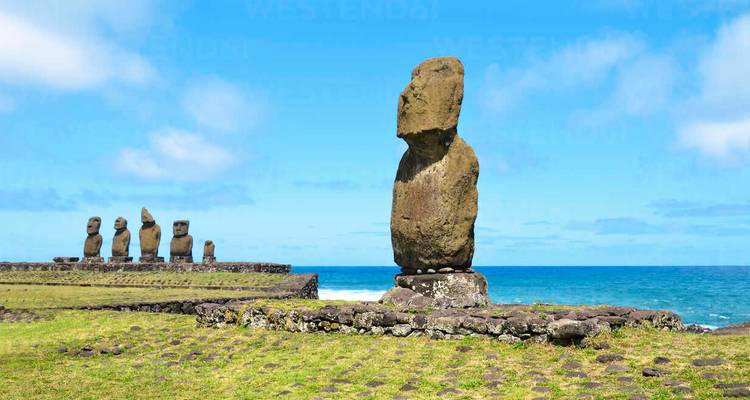 Estatuas Moai alineadas a lo largo de una plataforma de roca volcánica cubierta de hierba bajo un cielo azul brillante y el horizonte del océano.