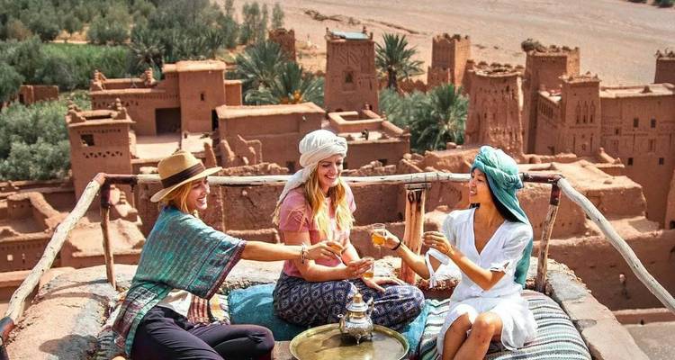 Trois jeunes femmes en foulards traditionnels portent un toast au thé sur un toit surplombant une ancienne kasbah.