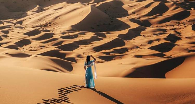 Figure en robe bleue flottante se tenant seule parmi de vastes dunes dorées laissant une traînée d'empreintes de pas.