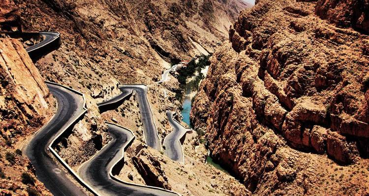 Une route goudronnée sinueuse serpente à travers les falaises rouges escarpées des gorges du Dadès avec un éclat de rivière en contrebas.