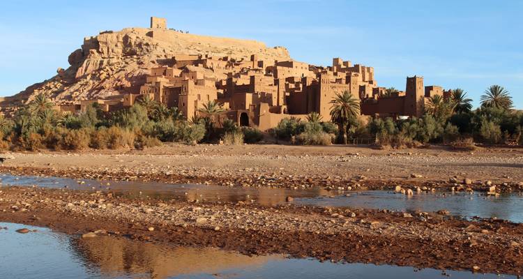 Le village fortifié en terre d'Aït Benhaddou s'élève au bord d'une rivière peu profonde sous un ciel bleu dégagé.