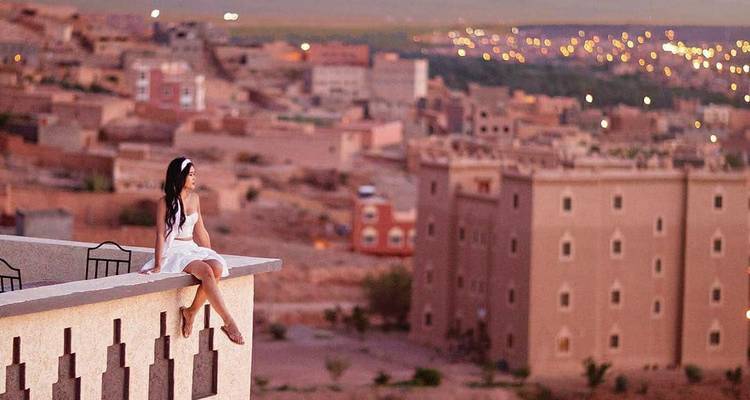 Femme en robe blanche assise sur le rebord d'un toit surplombant une ville marocaine aux tons terreux au crépuscule.