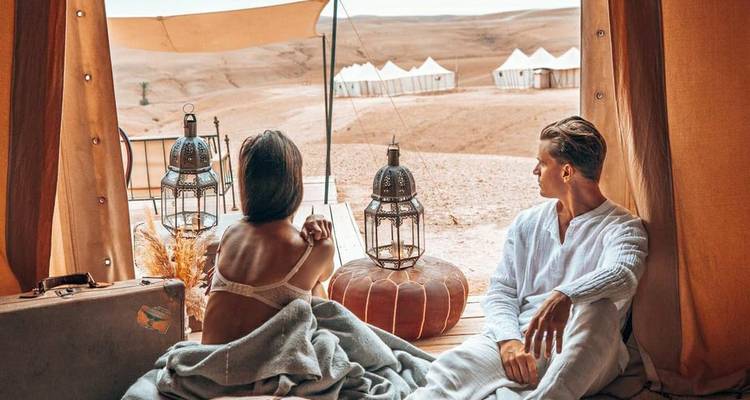Un couple se détend à l'intérieur d'une tente du désert haut de gamme en contemplant les dunes ondulantes et les tentes blanches du campement au loin.